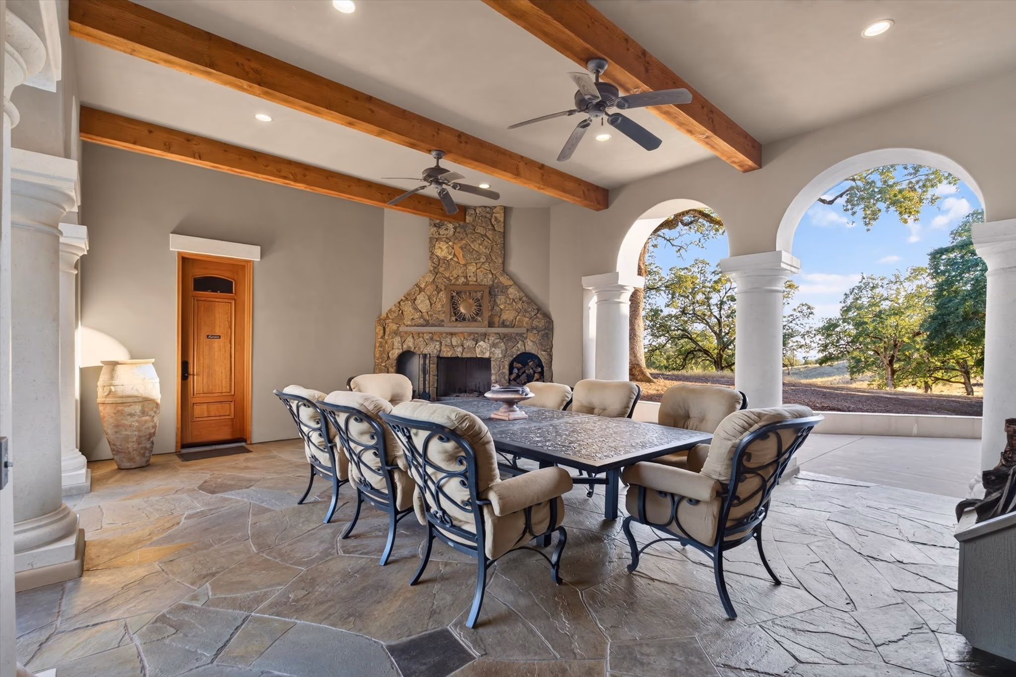 The covered outdoor dining loggia, with a beamed ceiling, a stone fireplace and a long table under arches open to the grounds.