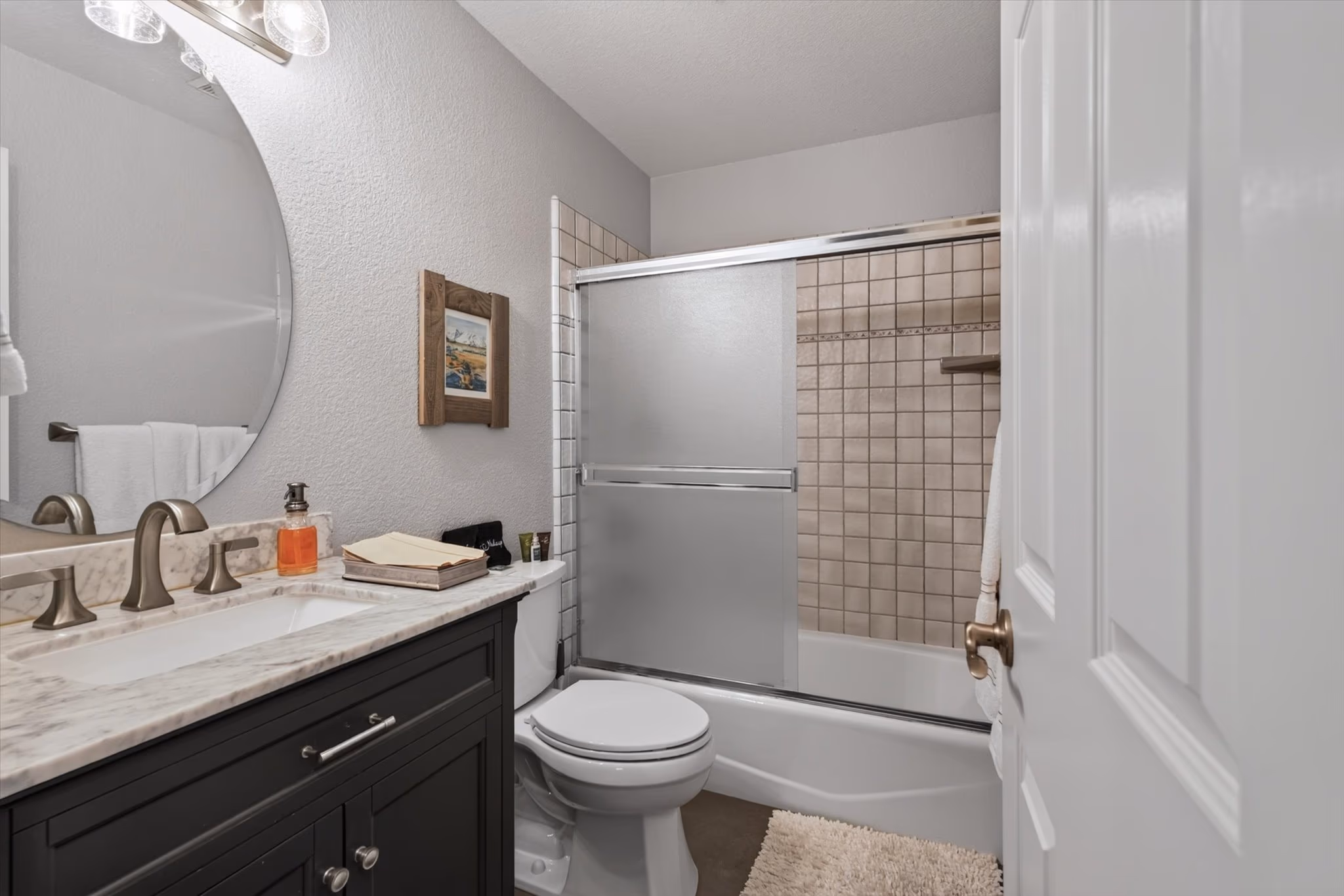A bathroom in the lake house, with a stone vanity, a round mirror and a tiled tub surround.