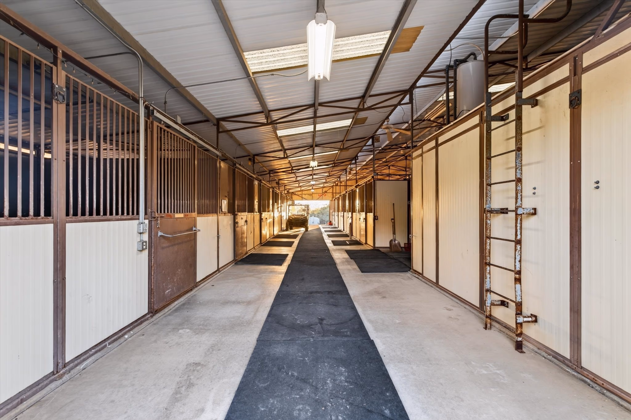 The interior aisle of the mare motel, lined with box stalls and lit by pendant fixtures, running to open doors at the far end.