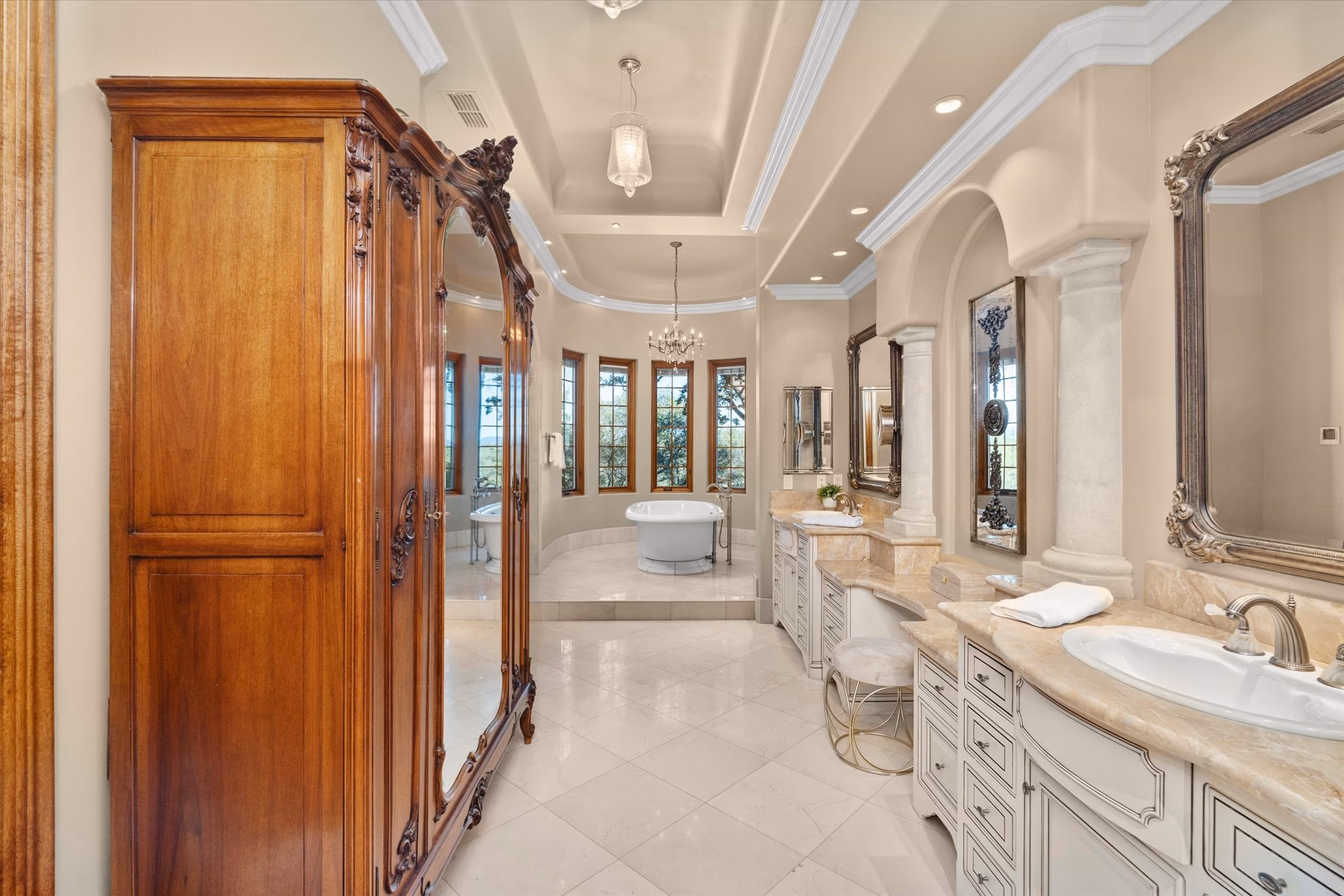 The primary bathroom, with twin vanities, stone columns, a carved armoire and the roll-top bath at the far end.