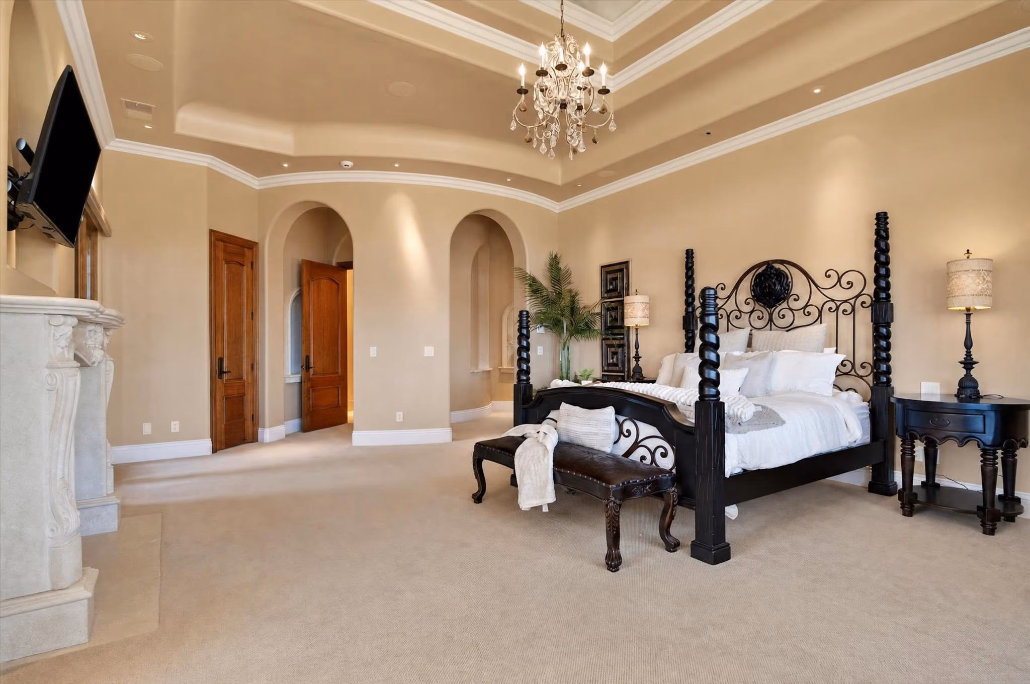 The primary bedroom, with a four-poster iron bed, a tray ceiling, a chandelier and arched openings to the bath beyond.