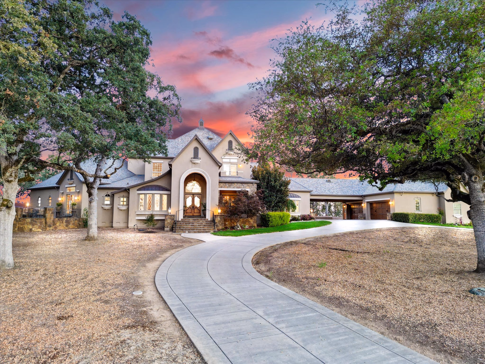 The French Country main residence at twilight, its arched entry and windows lit, framed by mature oaks with the circular drive in the foreground.